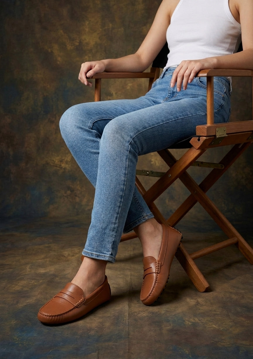 Person wearing tan brown loafers and blue jeans sitting on a wooden chair against a textured brown background